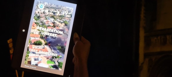 A person holds a tablet displaying an aerial view of a city with visible smoke and destruction. The screen includes text saying 'Devastation. Unrest. Free Palestine.' along with an emblem and hashtags.