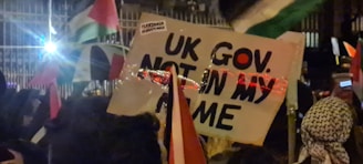 A group of people are gathered, holding various signs and flags. One prominent sign has the text 'UK GOV. NOT IN MY NAME'. The scene appears to be at nighttime or in low light with artificial lights visible in the background, creating reflections on some objects.