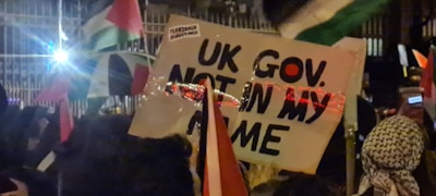 A group of people are gathered, holding various signs and flags. One prominent sign has the text 'UK GOV. NOT IN MY NAME'. The scene appears to be at nighttime or in low light with artificial lights visible in the background, creating reflections on some objects.