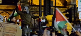 A crowded scene featuring people participating in a protest or demonstration. Some individuals are holding megaphones and flags, likely signifying a political or social cause. Various signs and placards are visible, with one individual prominently standing above the crowd. The background includes part of a building with illuminated windows, indicating a nighttime setting.