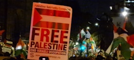 A nighttime scene featuring a political protest. A large sign with the message 'Free Palestine, End Israeli Occupation' is prominently displayed. The sign includes a graphic of the Palestinian flag and smaller text references to the Palestine Solidarity Campaign. A crowd of protesters is in the background, with many waving flags and holding various signs.