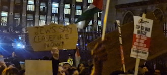 A crowd of people is gathered in a nighttime protest outside a building with many lit windows. They are holding cardboard signs with messages, one of which reads 'Jews Say Ceasefire Now!' and another says 'Stop the War on Gaza.' The atmosphere is charged and the scene is illuminated by artificial light.