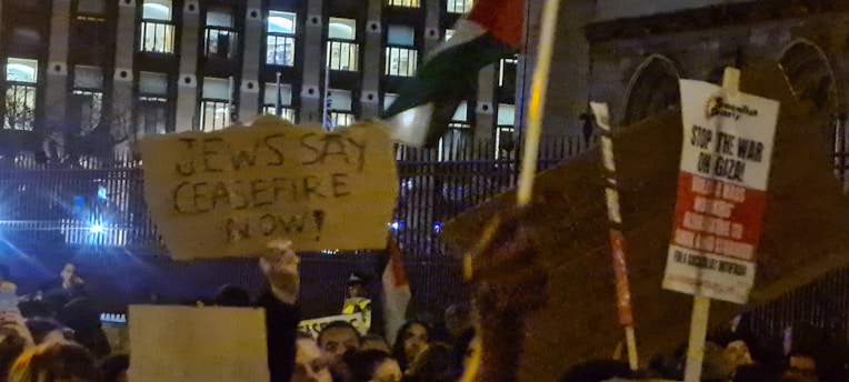 A crowd of people is gathered in a nighttime protest outside a building with many lit windows. They are holding cardboard signs with messages, one of which reads 'Jews Say Ceasefire Now!' and another says 'Stop the War on Gaza.' The atmosphere is charged and the scene is illuminated by artificial light.