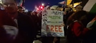 A nighttime gathering of people holding a sign with the text 'Palestine will be free' adorned with decorative leaves and waves. The crowd is surrounded by flags, and there are bright city lights and buildings visible in the background.