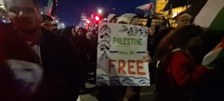 A nighttime gathering of people holding a sign with the text 'Palestine will be free' adorned with decorative leaves and waves. The crowd is surrounded by flags, and there are bright city lights and buildings visible in the background.