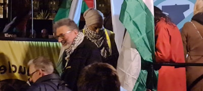 Several people are gathered in a dimly lit setting, featuring flags with green, white, red, and black colors. One person is smiling and wearing a scarf with a black and white pattern. The background includes a yellow banner with partial text and a nighttime atmosphere.