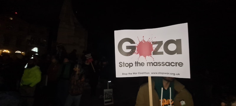 A protestor holds a sign that reads 'Gaza Stop the massacre' during a night protest. The crowd surrounding the individual is partially visible, with some people carrying signs and wearing warm clothes. The setting appears dark, suggesting the event is taking place at night.