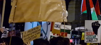 A crowd of people holding various signs and banners, some bearing messages of political activism and support for Palestine. There are flags in the background, and the scene appears to be at night or in a dimly lit area.