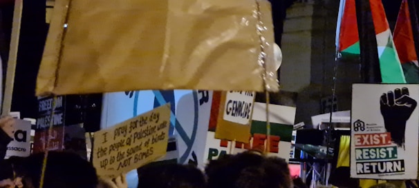 A crowd of people holding various signs and banners, some bearing messages of political activism and support for Palestine. There are flags in the background, and the scene appears to be at night or in a dimly lit area.