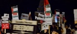 A group of people are holding various protest signs advocating for Palestinian freedom. The signs feature messages such as 'Free Palestine', 'Freedom for Palestine', and other similar statements. The crowd appears to be gathered in a demonstration or rally.