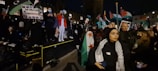 A group of people gathered at night, many of them holding flags and protest signs. Some individuals appear to be standing on a platform, with one person using a megaphone. The crowd seems to be actively participating, and there are several national flags visible.