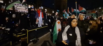 A group of people gathered at night, many of them holding flags and protest signs. Some individuals appear to be standing on a platform, with one person using a megaphone. The crowd seems to be actively participating, and there are several national flags visible.