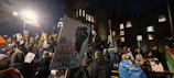 A group of people is gathered during nighttime for what appears to be a protest or demonstration. Some individuals are holding flags and placards. One prominent sign displays a fist and has the words 'EXIST. RESIST. RETURN'. The event seems to be taking place in a historic or significant location with a large, illuminated building in the background.