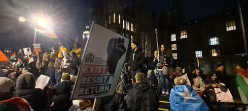 A group of people is gathered during nighttime for what appears to be a protest or demonstration. Some individuals are holding flags and placards. One prominent sign displays a fist and has the words 'EXIST. RESIST. RETURN'. The event seems to be taking place in a historic or significant location with a large, illuminated building in the background.
