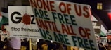 A protest scene with various signs and placards, one prominently displaying the message 'None of us are free until all of us are free.' Another sign references Gaza and stopping the massacre. The crowd is holding multiple banners with political and activist messages.