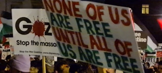 A protest scene with various signs and placards, one prominently displaying the message 'None of us are free until all of us are free.' Another sign references Gaza and stopping the massacre. The crowd is holding multiple banners with political and activist messages.