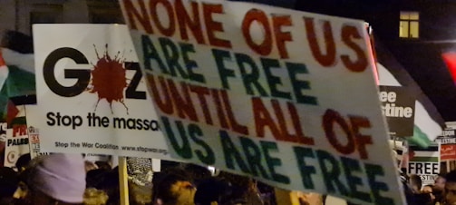 A protest scene with various signs and placards, one prominently displaying the message 'None of us are free until all of us are free.' Another sign references Gaza and stopping the massacre. The crowd is holding multiple banners with political and activist messages.