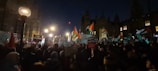A nighttime protest with a large crowd holding flags and placards. The crowd is gathered in front of a historic building with illuminated windows. The area is lit by street lamps, and various signs are visible, including those with messages of freedom.