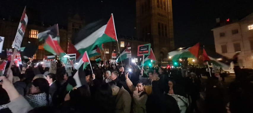 A large group of people are gathered in a nighttime setting holding flags and signs in what appears to be a demonstration. The flags are prominently red, white, green, and black, suggesting a particular national identity. The background includes a tall, well-lit building, possibly of historic or governmental significance.