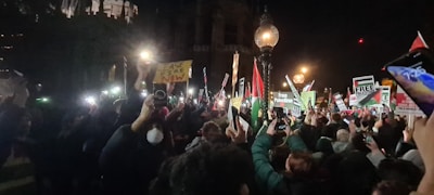 A large group of people are gathered at night holding up signs and placards, many of which have messages related to a political demonstration. The crowd is dense, with people raising their hands and phones, capturing the event. Street lamps illuminate the scene, adding a warm glow.