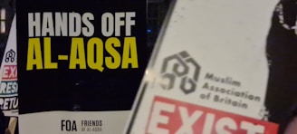 A protest sign displays bold yellow and white text against a black background reading 'Hands Off Al-Aqsa.' Nearby, another sign with the logo of the Muslim Association of Britain emphasizes its message.
