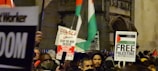 A crowd is gathered, holding various signs and flags related to political activism. Some signs include text advocating for Palestine, with phrases such as 'Free Palestine' and 'Stop the War on Gaza'. People in the crowd are dressed warmly, suggesting a nighttime or cool-weather setting.