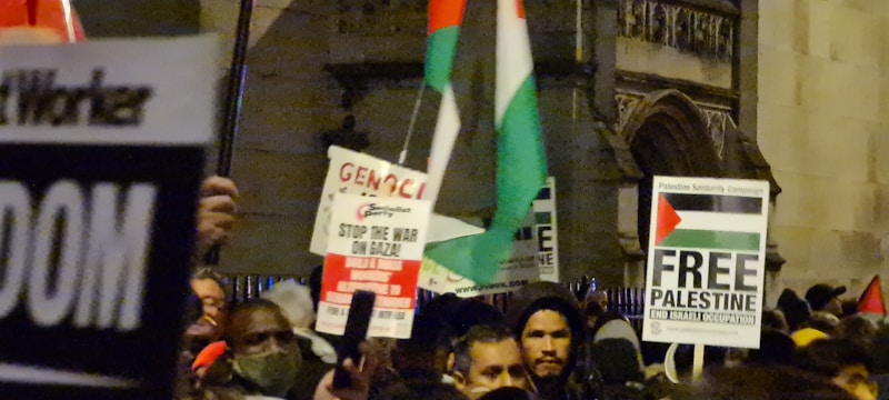 A crowd is gathered, holding various signs and flags related to political activism. Some signs include text advocating for Palestine, with phrases such as 'Free Palestine' and 'Stop the War on Gaza'. People in the crowd are dressed warmly, suggesting a nighttime or cool-weather setting.