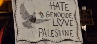 A sign with a hand-drawn image of a dove carrying a flag. The text on the sign reads 'Hate Genocide, Love Palestine' with a heart symbol included.
