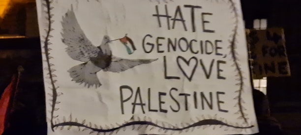 A sign with a hand-drawn image of a dove carrying a flag. The text on the sign reads 'Hate Genocide, Love Palestine' with a heart symbol included.