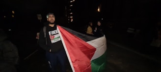 A person stands while holding a large flag with the design and colors of the Palestinian flag. The individual is wearing a sweatshirt with text related to Palestine, and the setting appears to be outdoors at night with limited lighting.