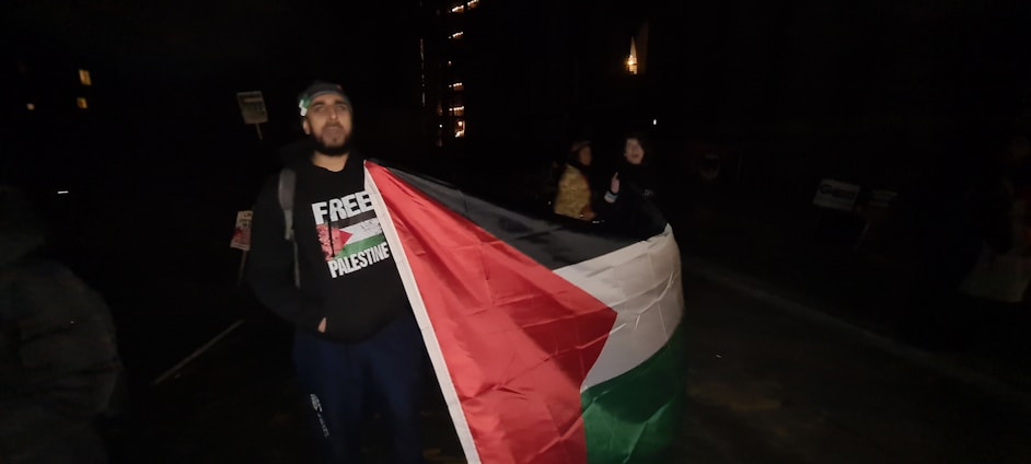 A person stands while holding a large flag with the design and colors of the Palestinian flag. The individual is wearing a sweatshirt with text related to Palestine, and the setting appears to be outdoors at night with limited lighting.