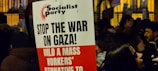 A protest sign with the heading 'Socialist Party' and messages including 'Stop the War on Gaza!' and 'Build a Mass Workers’ Alternative to'. The sign is held in a crowded area where people are gathered, possibly during a demonstration. The background shows more signs and people, partially obscured and dimly lit, suggesting a nighttime setting.