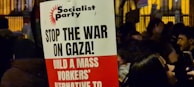 A protest sign with the heading 'Socialist Party' and messages including 'Stop the War on Gaza!' and 'Build a Mass Workers’ Alternative to'. The sign is held in a crowded area where people are gathered, possibly during a demonstration. The background shows more signs and people, partially obscured and dimly lit, suggesting a nighttime setting.