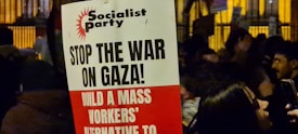 A protest sign with the heading 'Socialist Party' and messages including 'Stop the War on Gaza!' and 'Build a Mass Workers’ Alternative to'. The sign is held in a crowded area where people are gathered, possibly during a demonstration. The background shows more signs and people, partially obscured and dimly lit, suggesting a nighttime setting.