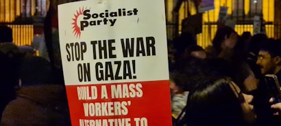A protest sign with the heading 'Socialist Party' and messages including 'Stop the War on Gaza!' and 'Build a Mass Workers&rsquo; Alternative to'. The sign is held in a crowded area where people are gathered, possibly during a demonstration. The background shows more signs and people, partially obscured and dimly lit, suggesting a nighttime setting.