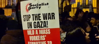 A protest sign with the heading 'Socialist Party' and messages including 'Stop the War on Gaza!' and 'Build a Mass Workers’ Alternative to'. The sign is held in a crowded area where people are gathered, possibly during a demonstration. The background shows more signs and people, partially obscured and dimly lit, suggesting a nighttime setting.