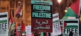 Protesters holding signs with messages supporting Palestinian freedom and solidarity, amidst a crowd waving flags.