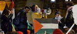 A group of people are gathered, with one person speaking into a megaphone. There is a large flag with a red, green, and black triangular pattern. The gathering appears to be a protest or rally with several individuals engaged.