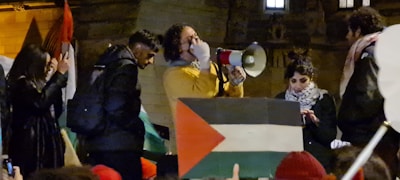 A group of people are gathered, with one person speaking into a megaphone. There is a large flag with a red, green, and black triangular pattern. The gathering appears to be a protest or rally with several individuals engaged.