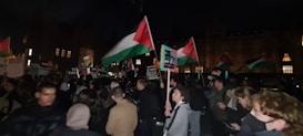 A nighttime gathering with a large group of people holding flags and signs. The flags are red, white, green, and black, and there are various placards raised above the crowd. The setting appears to be outside a large building with illuminated windows.