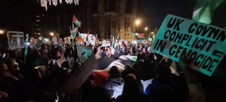 A large group of people gathered in a nighttime protest, holding flags and signs with various messages. The scene is illuminated by streetlights, and the crowd appears focused and engaged.