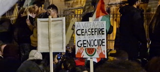 A crowd of people gathered at night, some holding signs with messages. The main sign is in focus with text stating 'A vote against ceasefire is a vote for genocide' along with imagery of hands and a watermelon.