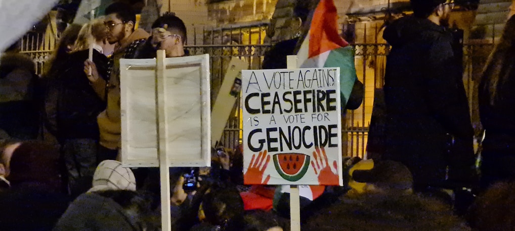 A crowd of people gathered at night, some holding signs with messages. The main sign is in focus with text stating 'A vote against ceasefire is a vote for genocide' along with imagery of hands and a watermelon.