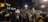 A large group of people gathered outdoors during the night, many of whom are holding various signs and banners. Some of the signs read 'Free Palestine' and 'Gaza, stop the massacre.' The crowd is densely packed, with many individuals wearing winter clothing such as jackets and hats.