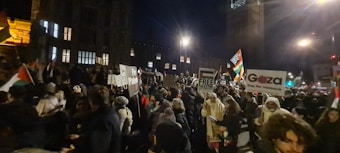 A large group of people gathered outdoors during the night, many of whom are holding various signs and banners. Some of the signs read 'Free Palestine' and 'Gaza, stop the massacre.' The crowd is densely packed, with many individuals wearing winter clothing such as jackets and hats.