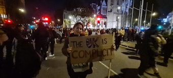 A person holding a protest sign that reads 'The UK is Complicit' stands amidst a large group of people gathered on a city street at night. The scene is illuminated by streetlights and nearby buildings, including a notable architectural structure in the background. The atmosphere appears to be part of a demonstration or protest.