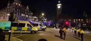 A nighttime scene featuring several police vans parked on a city street with a prominent clock tower in the background. Police officers in uniform are standing and walking around the area, while a group of people is gathered nearby. The surrounding buildings are lit, creating a contrast with the night sky.
