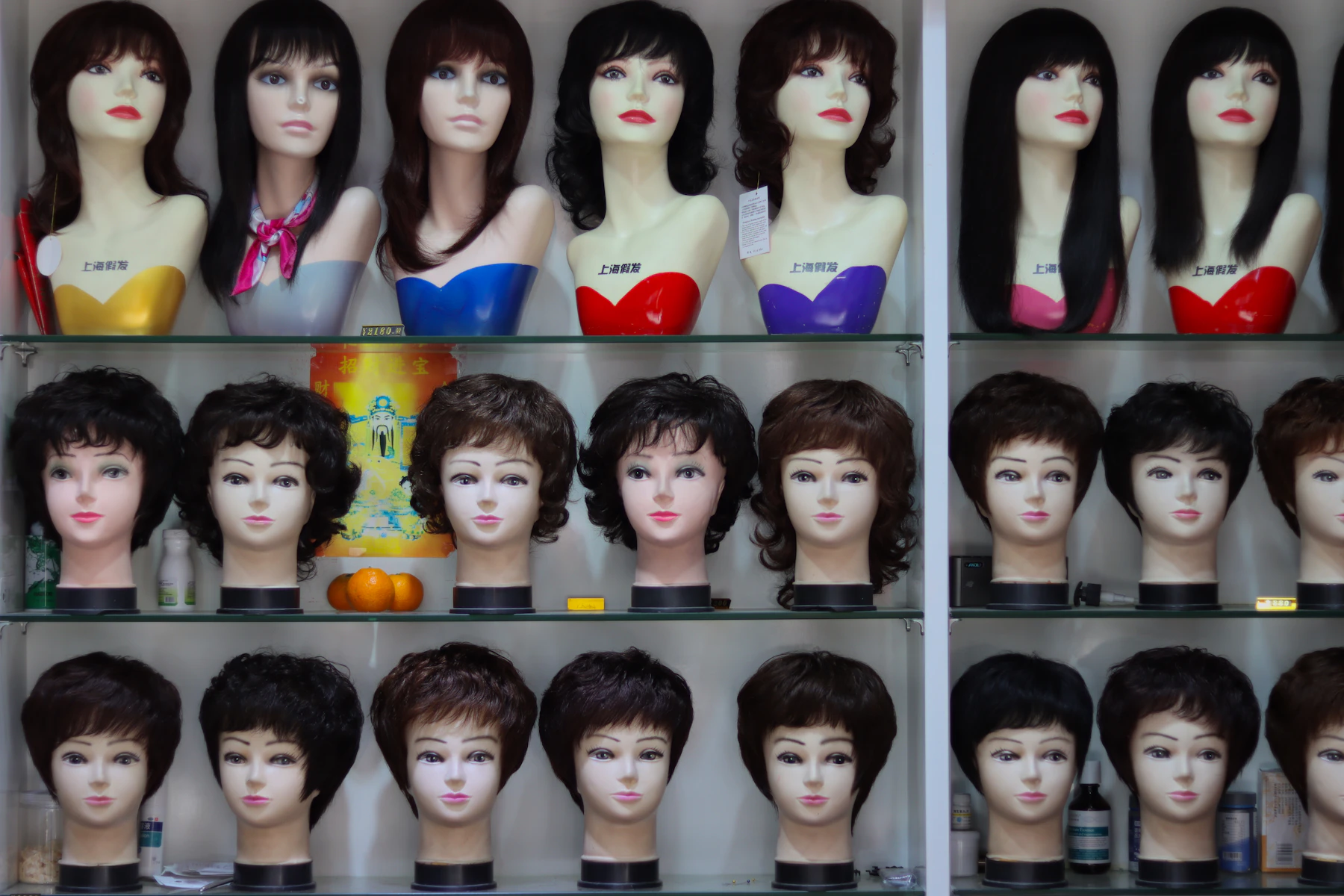 Glass shelves of mannequin heads wearing short and long dark wigs, three rows deep