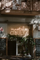 A cozy storefront features a sign with 'Vinotéka Cibulka' in glowing letters. The entrance is adorned with greenery, creating an inviting atmosphere. Chalkboards with writing are positioned on either side, listing various items or daily specials. The architecture includes a balcony with a decorative iron railing above.