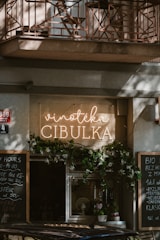 A cozy storefront features a sign with 'Vinotéka Cibulka' in glowing letters. The entrance is adorned with greenery, creating an inviting atmosphere. Chalkboards with writing are positioned on either side, listing various items or daily specials. The architecture includes a balcony with a decorative iron railing above.